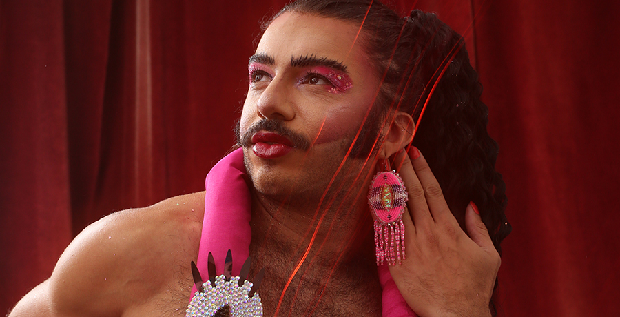 Alex, Glamputee, looks up and to the left side of the picture, pursing lips. Glamputee is in drag makeup, wearing bright pink eyeshadow, blush, and lipstick. They wears long, bright pink dangly beaded earrings, and have a bright pink decorative stuffed flamingo draped around their neck.