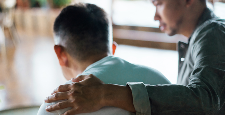 Rear view of son and elderly father sitting together at home. Son caring for his father, putting hand on his shoulder, comforting and consoling him. Family love, bonding, care and confidence