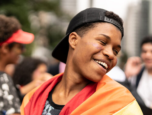 Young Black teenager outside surrounded by three other young people in the background; the boy in the foreground looks over his left shoulder, smiling, and is draped in a Pride flag over his shoulders.