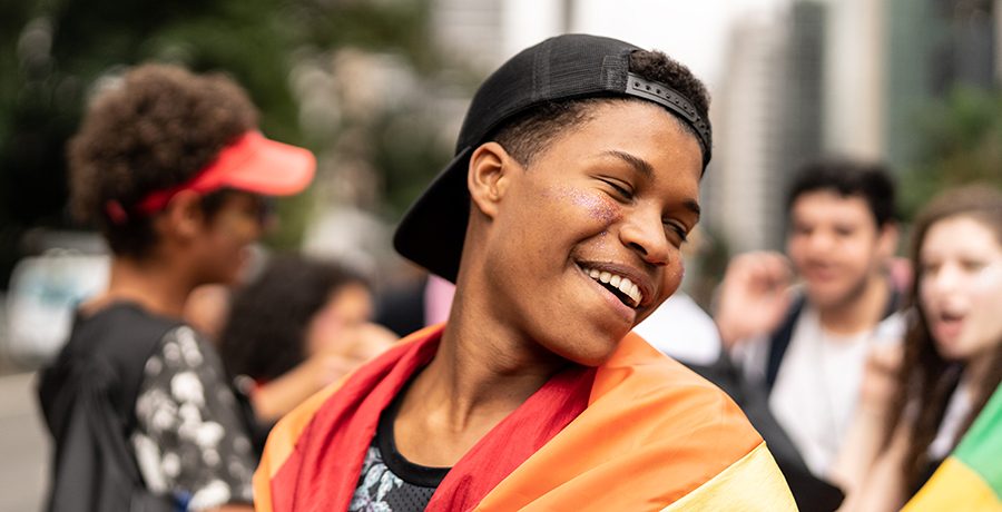 Young Black teenager outside surrounded by three other young people in the background; the boy in the foreground looks over his left shoulder, smiling, and is draped in a Pride flag over his shoulders.
