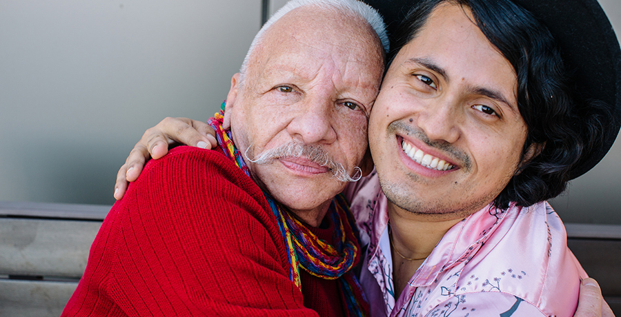 Two Latinx men face the camera, smiling. They have their arms around each other in a hug.